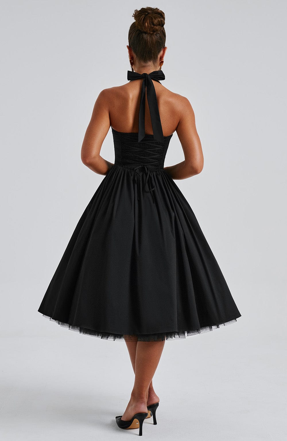 Back view of a black halter midi dress with a tied bow at the neck and a lace-up corset detail on the bodice, showing a full skirt with a tulle hem.