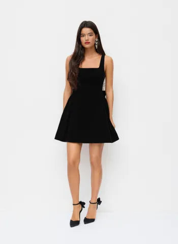 Full-length view of a woman wearing a black Lillie Bow Mini Dress, featuring a square neckline and bow detail on the back.