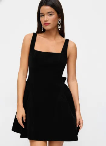 Mid-shot of a woman in a black Lillie Bow Mini Dress, highlighting the square neckline and subtle side bow accent.