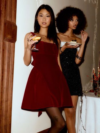 Model wearing a Lillie Bow Mini Dress in rich red velvet, holding a cocktail glass, with a subtle bow detail visible on the side.
