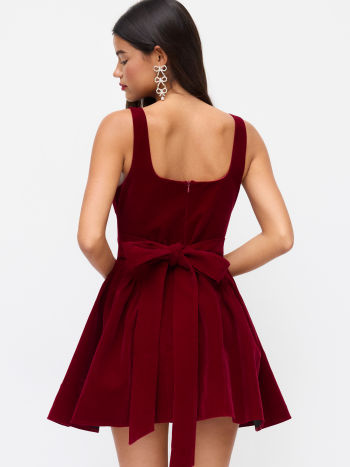 Rear view of a model in a Lillie Bow Mini Dress, made from red velvet, highlighting the square back, zipper closure, and large tied bow.