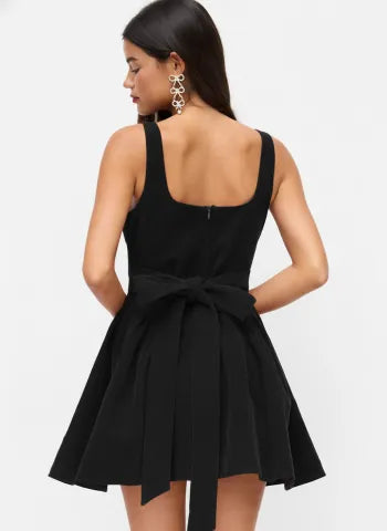 Rear view of a model wearing a Lillie Bow Mini Dress in black, showcasing the square back, subtle zipper, and a large tied bow at the waist.