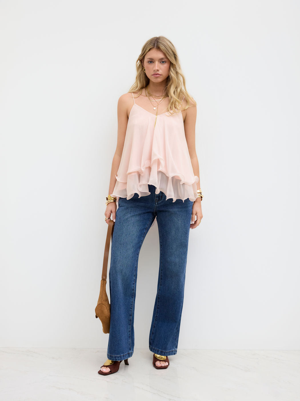 Blonde model standing, wearing a light pink Lily Ruffle Cami Top with layered ruffles, paired with dark wash jeans, gold jewelry, and brown heels.
