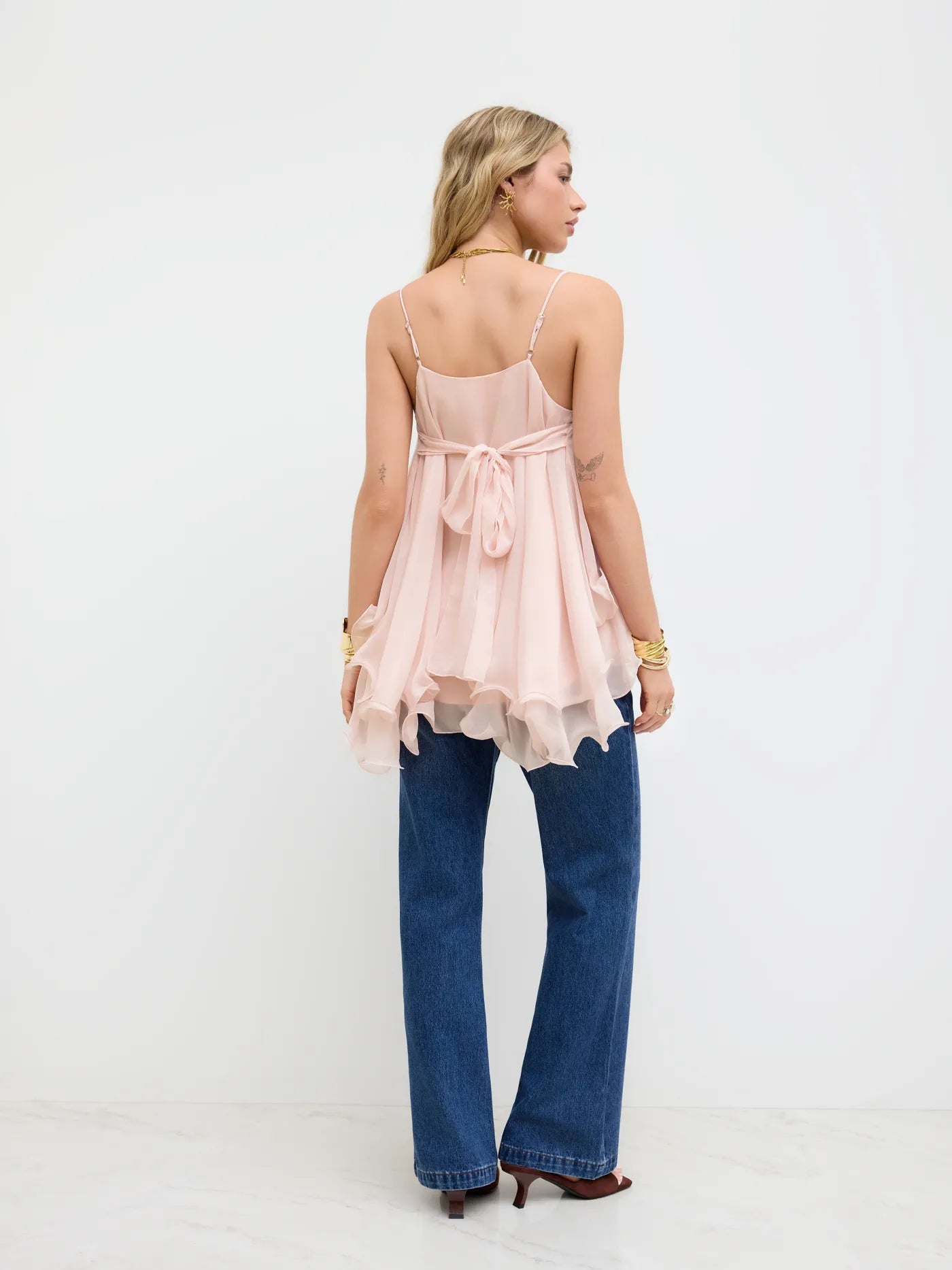 Back view of a blonde model wearing a light pink Lily Ruffle Cami Top, featuring a tie-back detail and showcasing the layered ruffles, paired with dark wash jeans.
