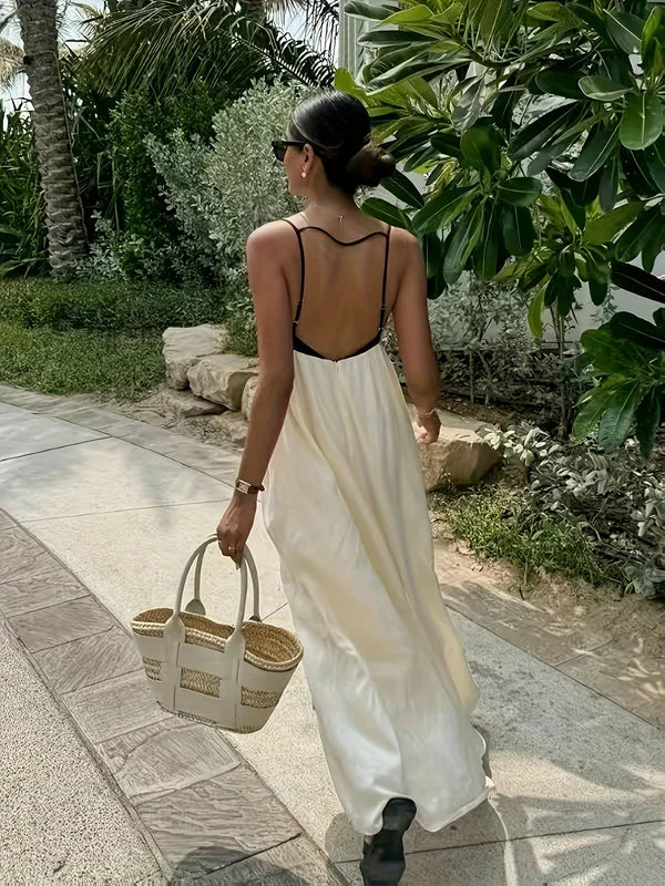 Back view of a woman wearing a cream and black color-block maxi dress with an open back and thin straps, walking outdoors.