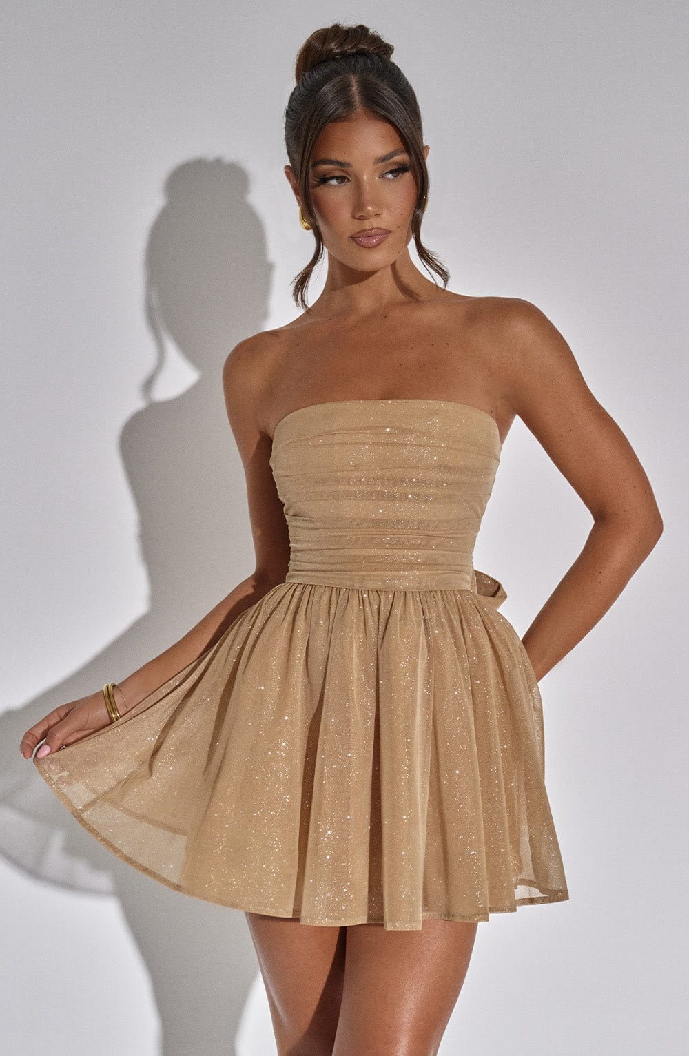 Champagne gold glitter mini dress with a strapless design and a flowing, pleated skirt, shown with one hand lifting the hem.