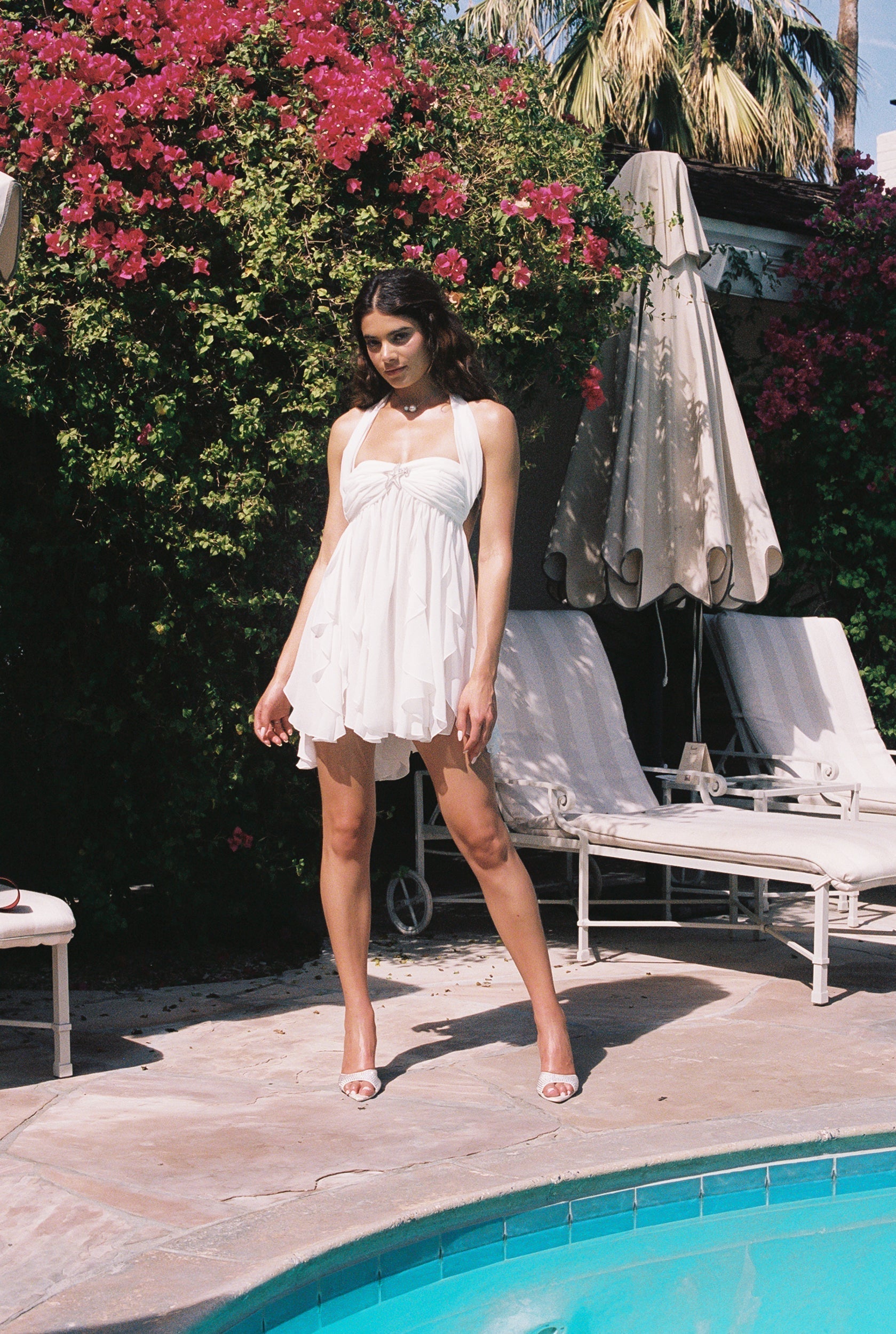 Full length view of a woman wearing a white halter neck mini dress with ruffled skirt, standing by a pool.