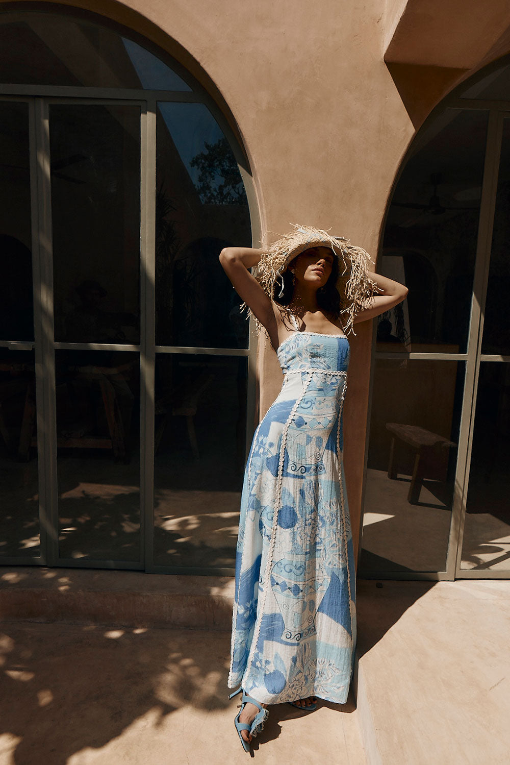 Woman wearing a straw hat and a blue and white strapless maxi dress, leaning against a wall in warm sunlight for women.