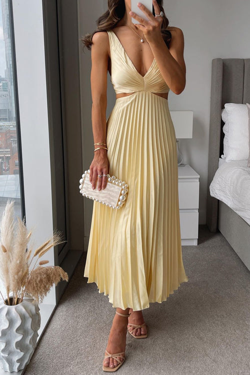 Maria Pleated Maxi Dress in light yellow with a deep V-neckline and side cut-outs, standing on a balcony overlooking the sea.