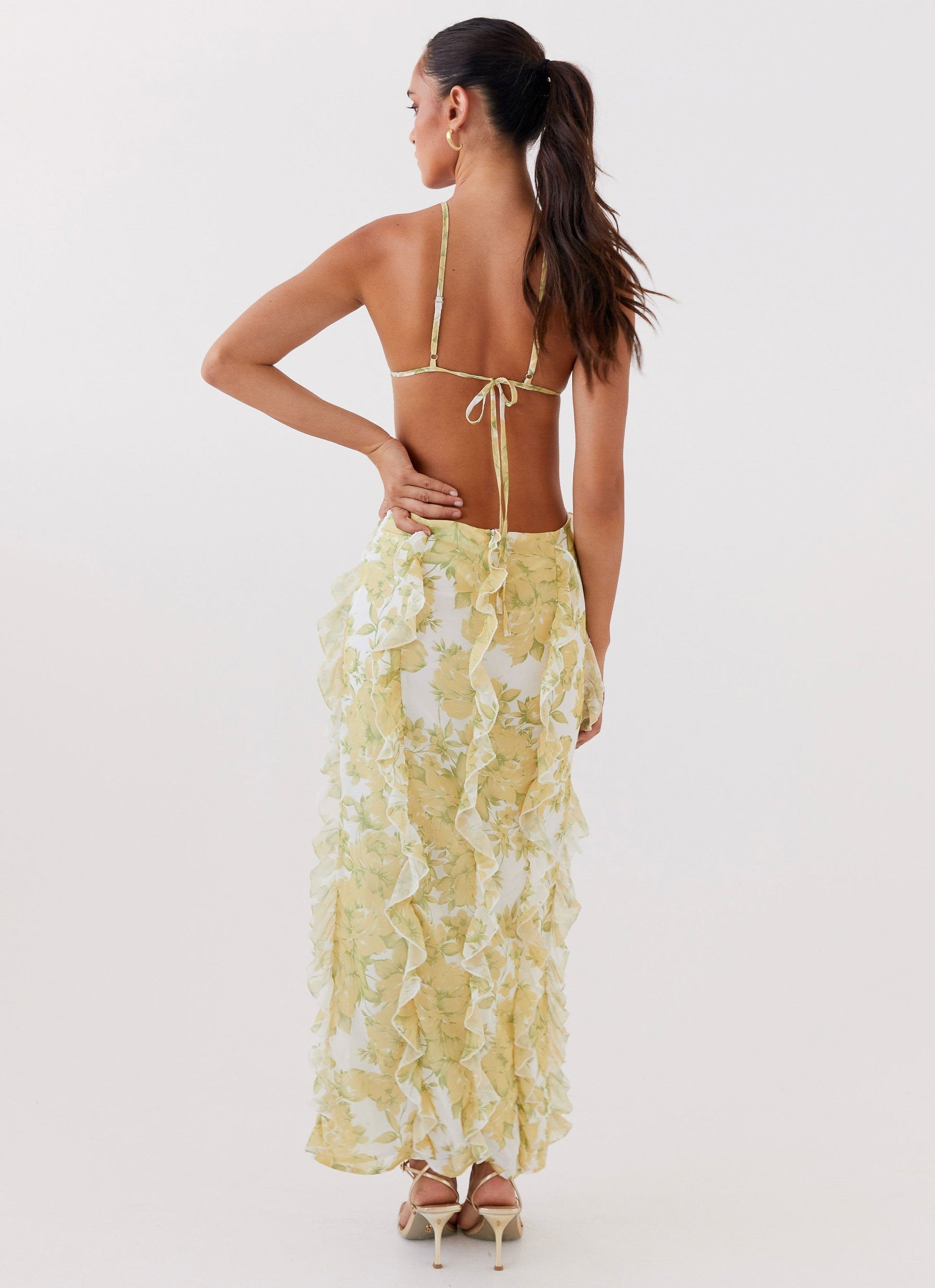 Back view of a woman in a white and yellow floral ruffled halter maxi dress with an open back tie closure