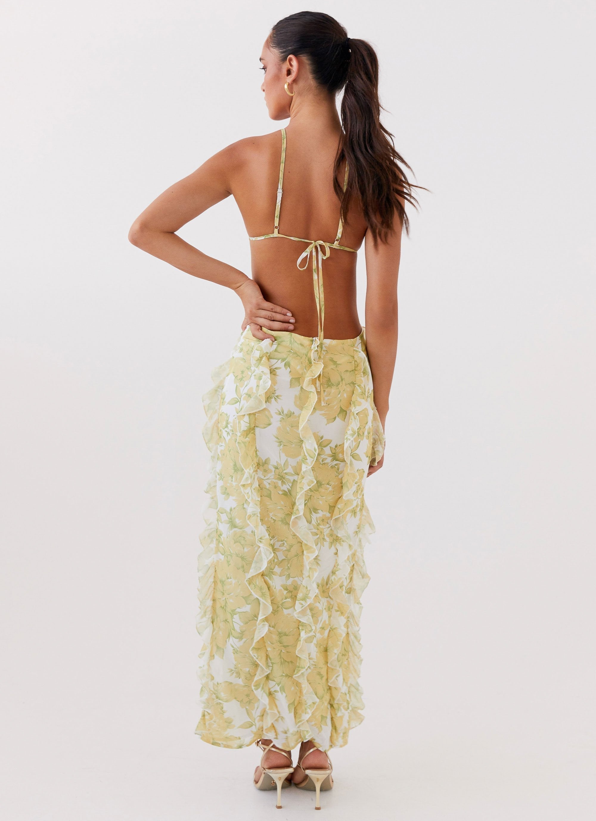 Back view of a woman in a white and yellow floral ruffled halter maxi dress with an open back tie closure
