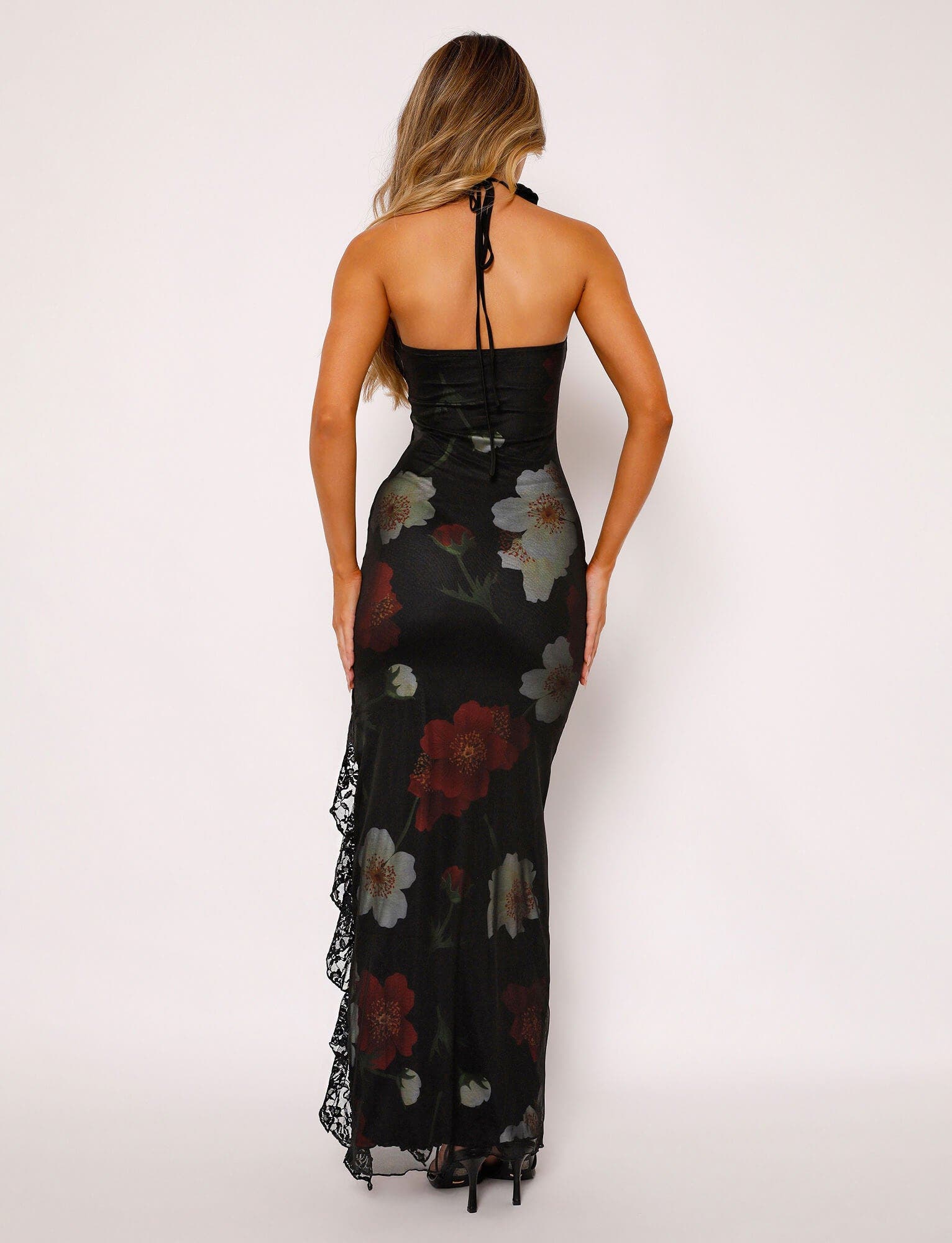 Back view of a black floral maxi dress with red and white flowers, halter neckline, and lace ruffle detail on the side.