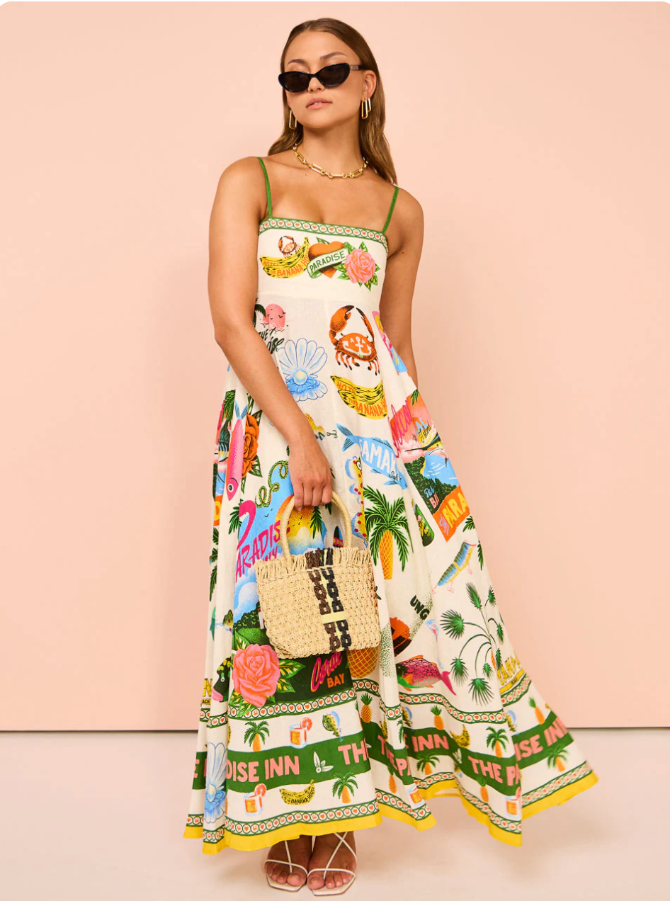 Full length view of a white tropical maxi dress with green spaghetti straps and colorful resort-themed print, worn with sunglasses and a straw bag.