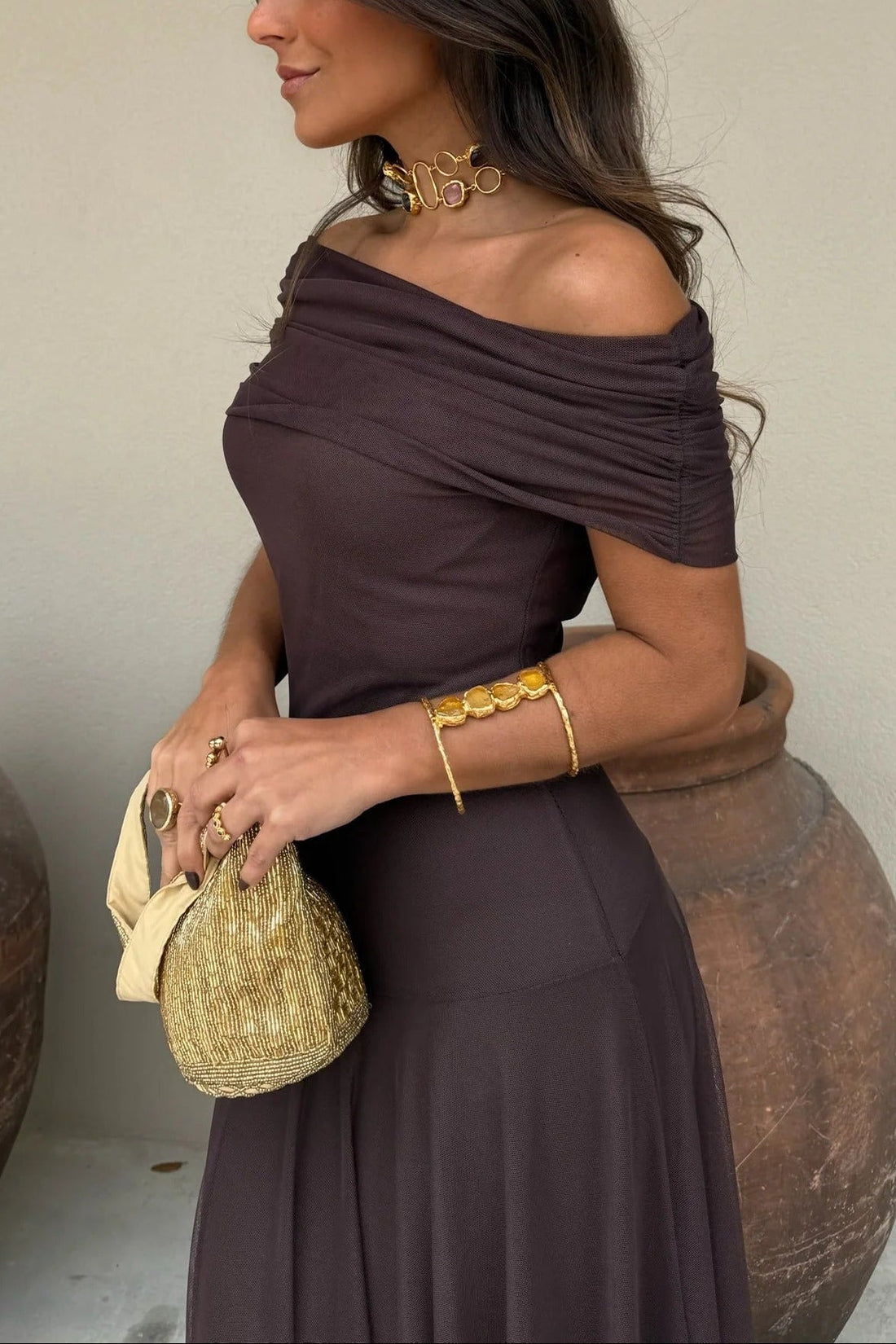Milena draped maxi dress in dark brown with one-shoulder design, flowy skirt, and gold accessories.