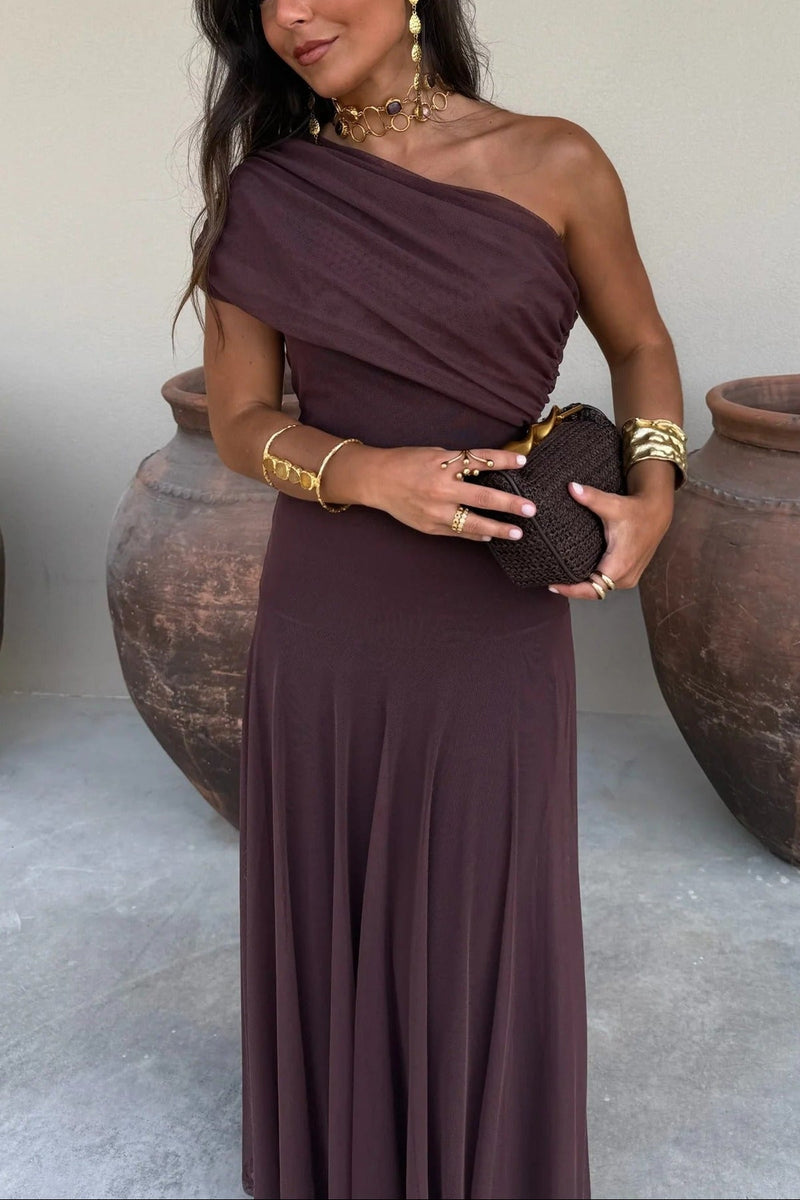 Milena draped maxi dress in dark brown with one-shoulder design, flowy skirt, and gold accessories.