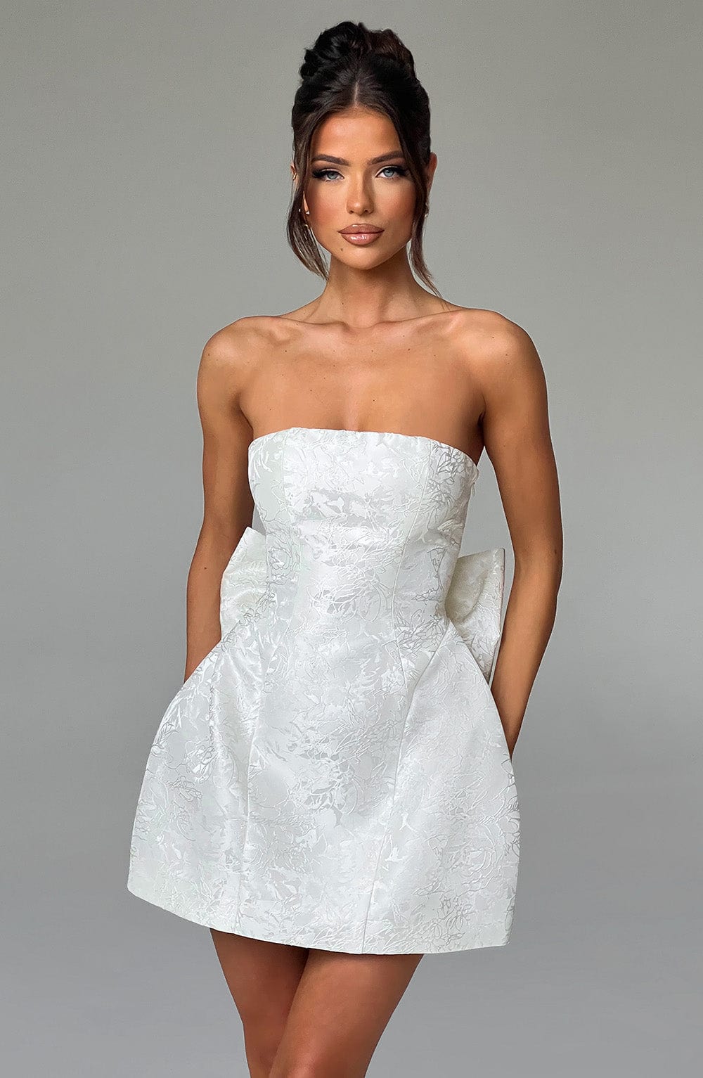 Front view of a white strapless mini dress with a textured floral pattern and a large bow on the side, a chic suit dress.