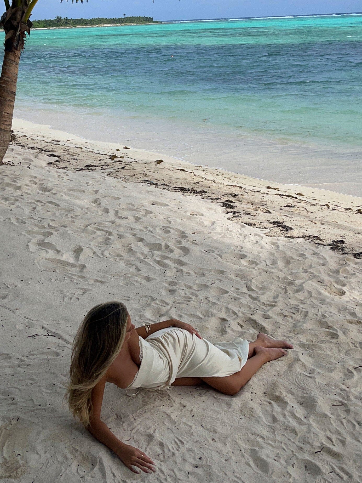 Full length shot of an ivory ruched maxi dress flowing on a woman laying on a sandy beach.