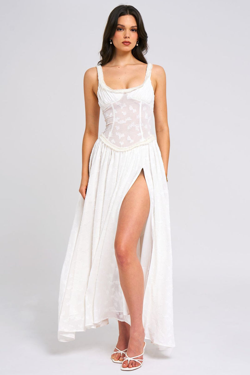 Elegant white Nina Corset Maxi Dress featuring a structured bodice, sheer paneling, and a high leg slit, worn with white strappy heels.