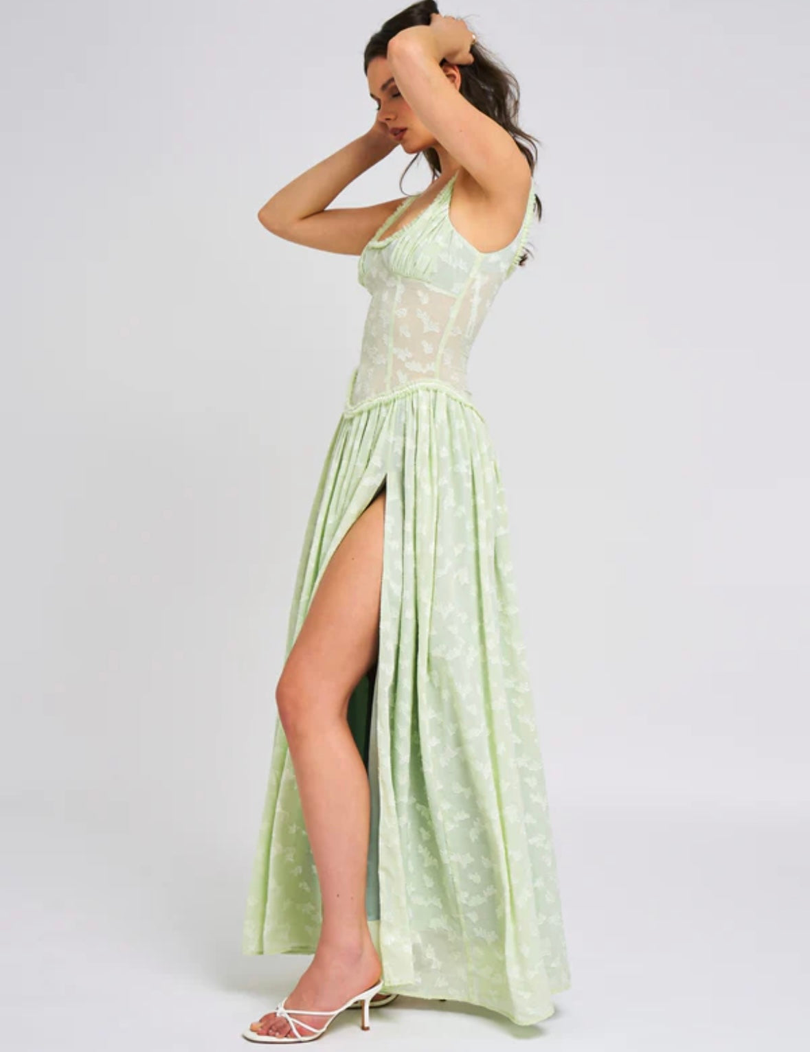 Side profile of the light green Nina Corset Maxi Dress, highlighting the thigh-high slit and the silhouette of the dress, with the model's hands in her hair.