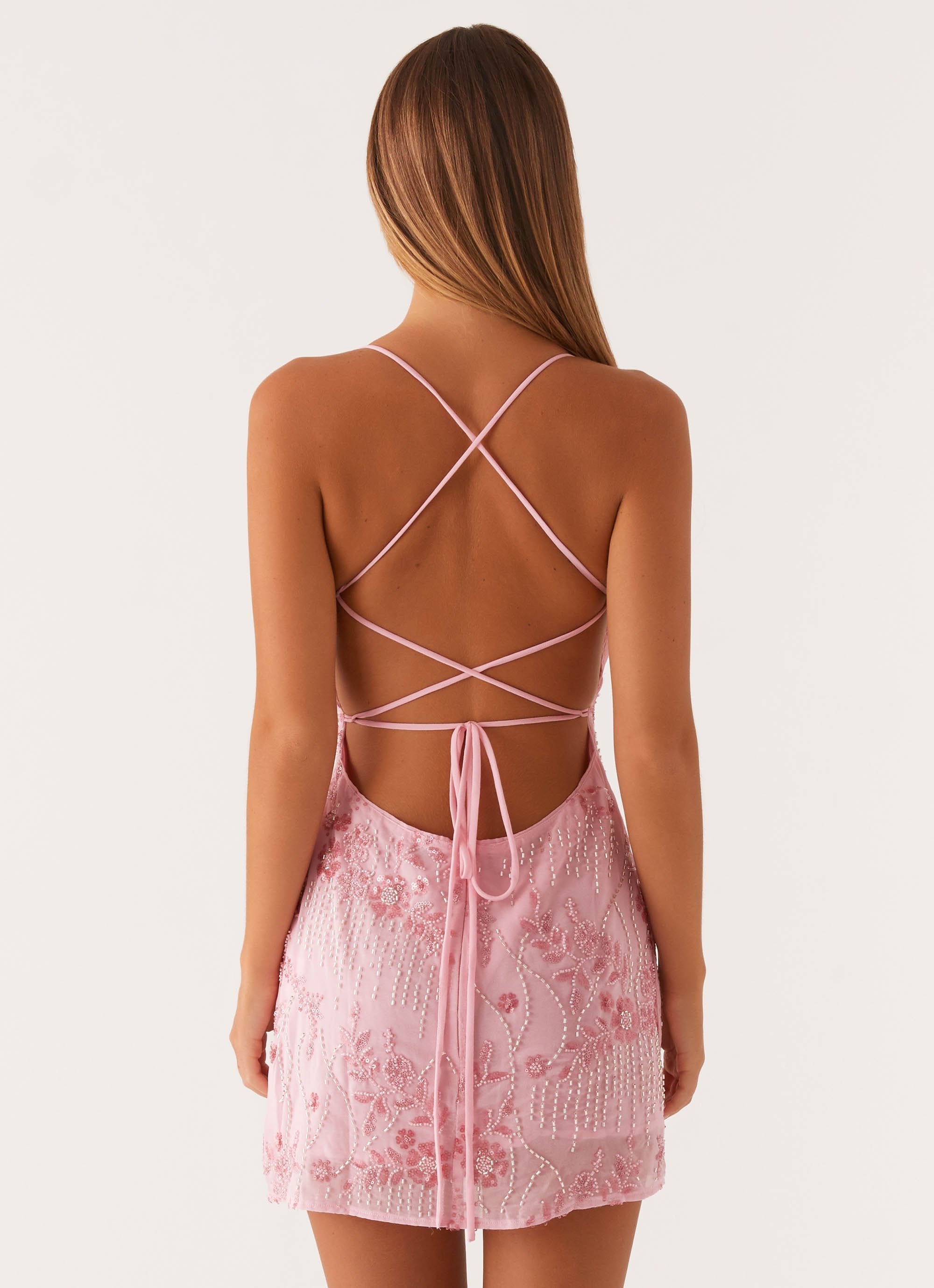 Back view of a pale pink bodycon mini dress, displaying the open back design with crisscross straps and a tie closure, accented with delicate floral and beaded patterns.
