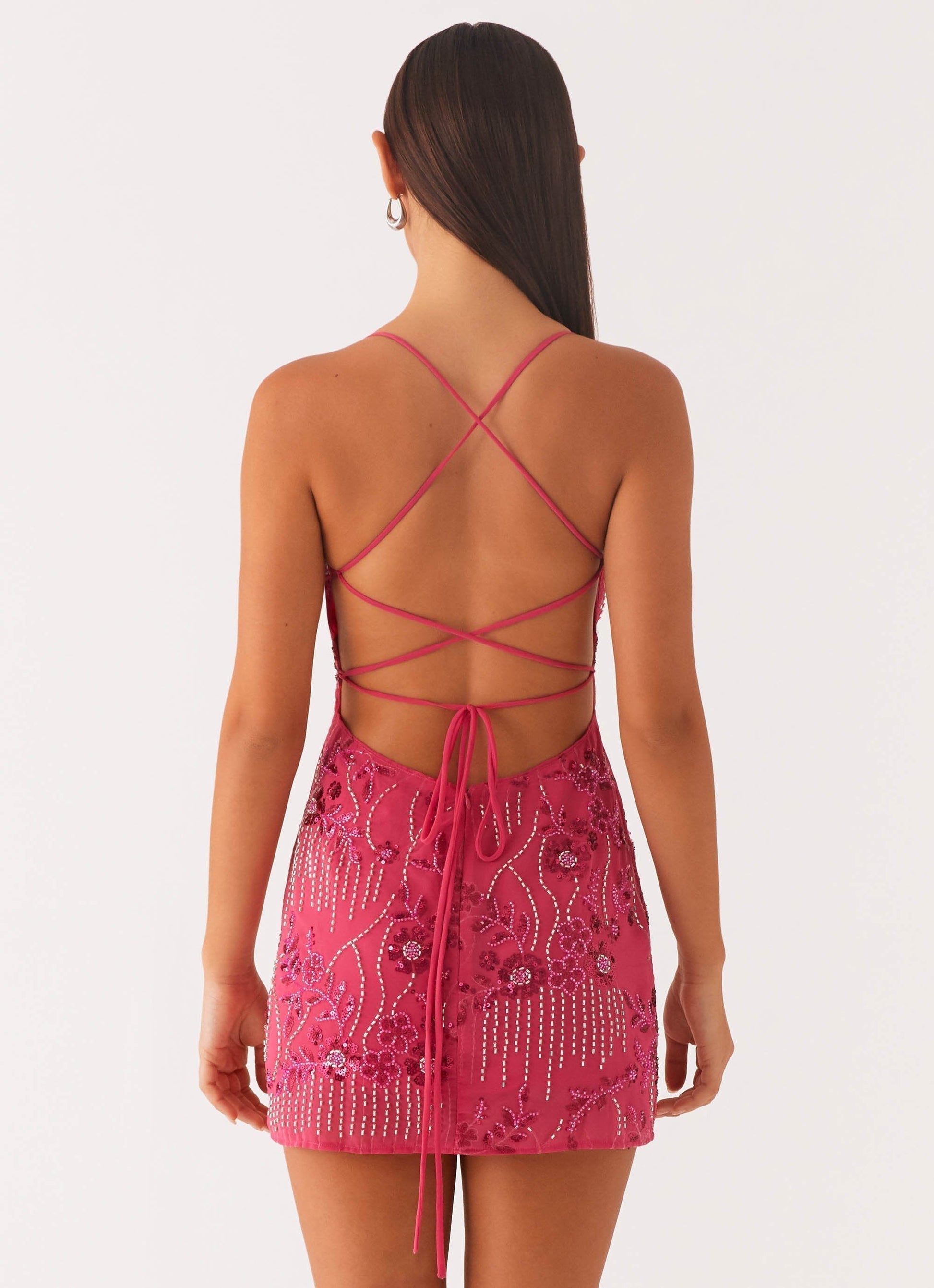 Back view of a fuchsia pink bodycon mini dress, highlighting the open back with crisscross spaghetti straps and a tie closure, adorned with floral and beaded patterns.