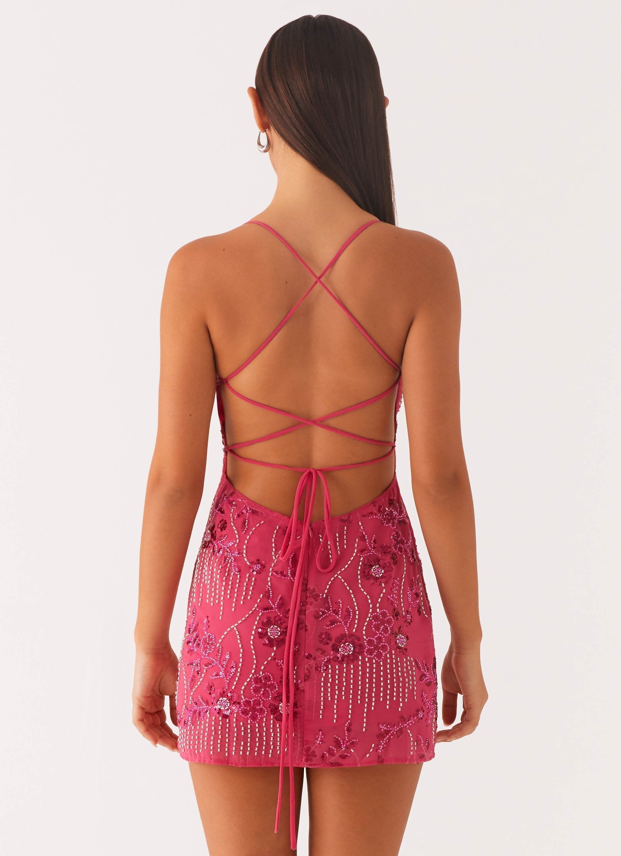 Back view of a fuchsia pink bodycon mini dress, highlighting the open back with crisscross spaghetti straps and a tie closure, adorned with floral and beaded patterns.