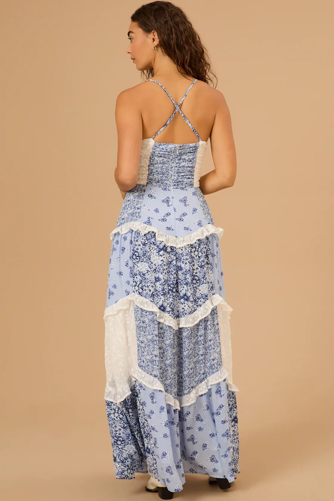 Rear view of the blue floral Nola Ruffle Maxi Dress, featuring the full tiered skirt, white lace inserts, and criss-cross back straps.