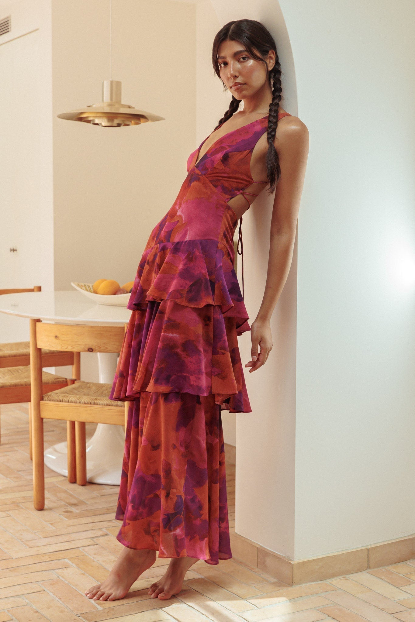 Side profile of a woman leaning against a wall, wearing a red and purple floral Harriet Ruffle Maxi Dress with tiered ruffles.