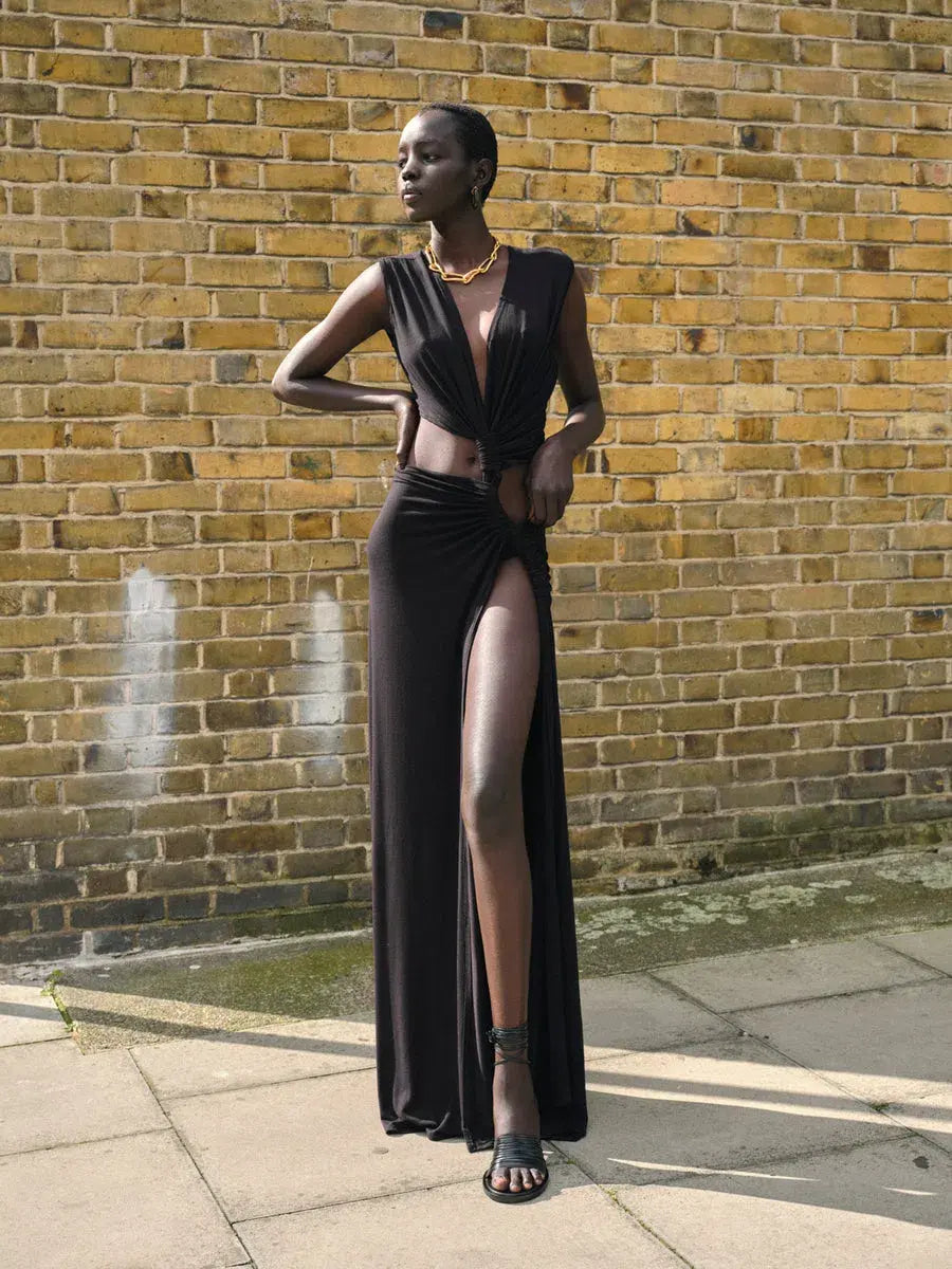 Black Robyn cutout maxi dress with a deep V-neck, side knot, and high slit, worn with black heels against a brick wall.