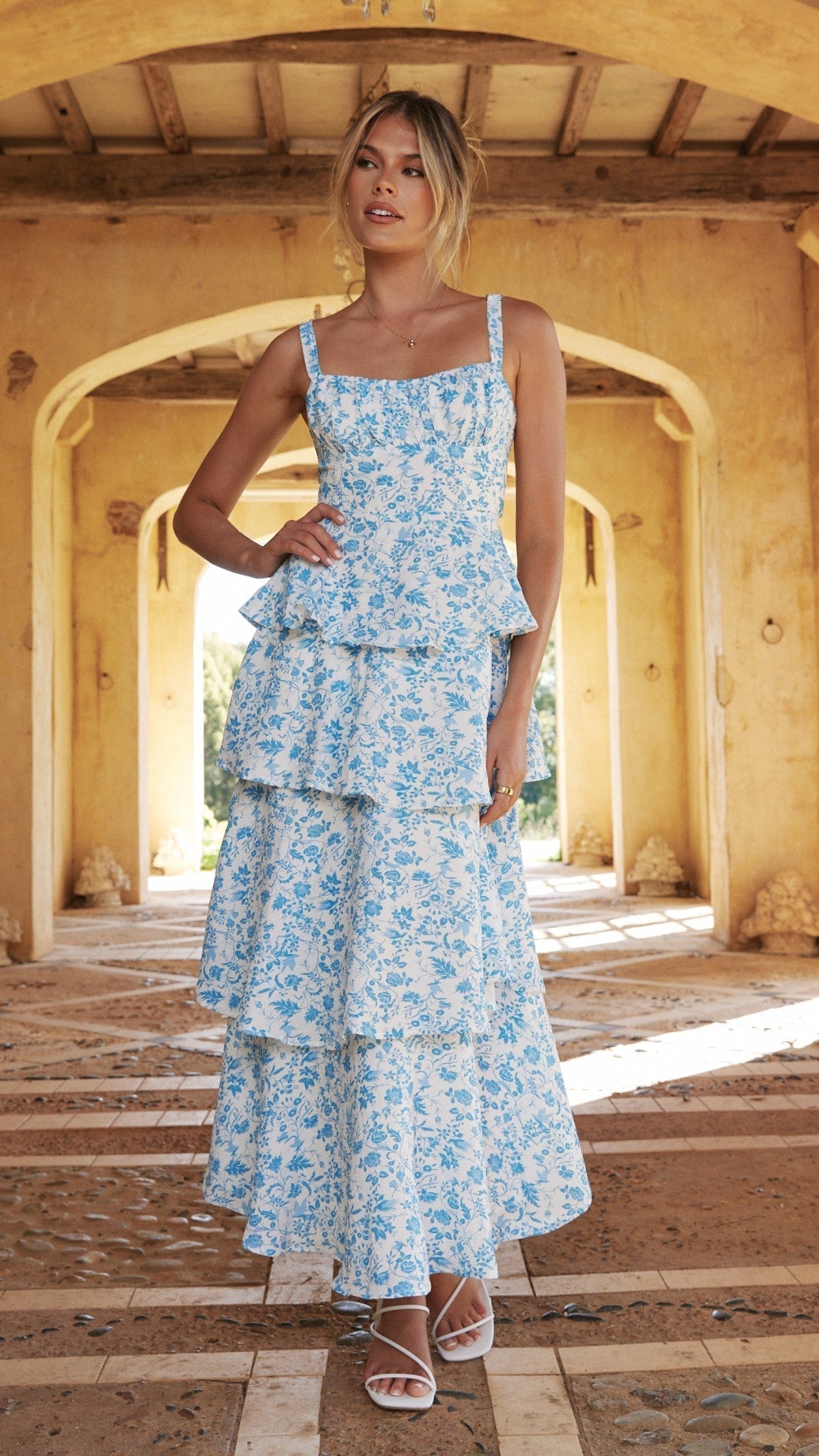 Rosanna Bow Maxi Dress in white with blue floral print, featuring a tiered skirt and peplum top, worn with white strappy sandals.