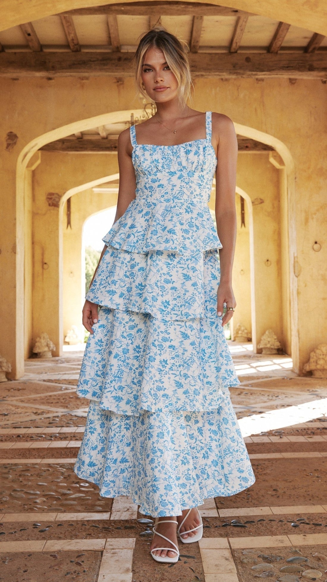 Front view of a white maxi dress with blue floral print, tiered skirt, and square neckline, worn with white heels.