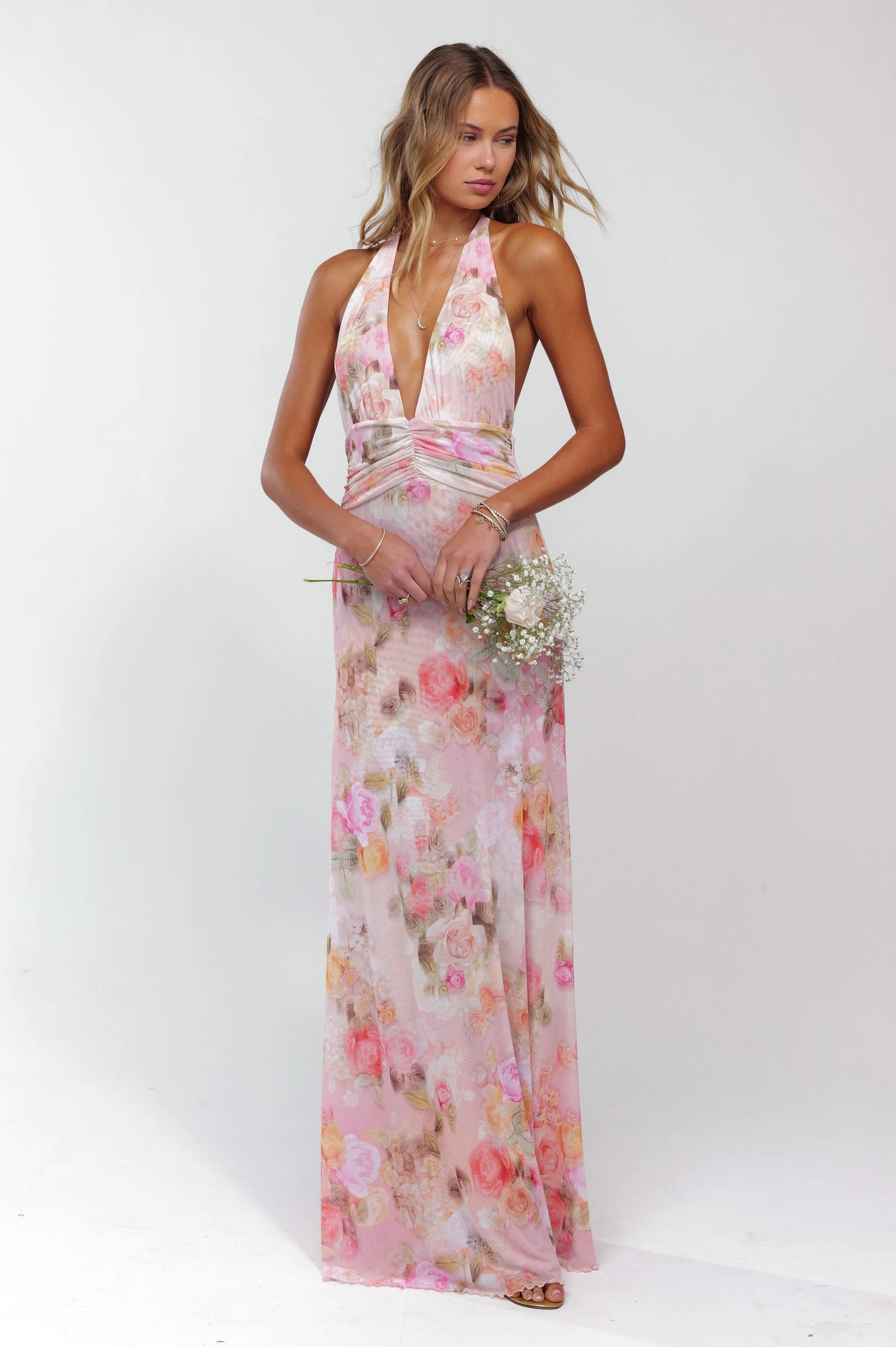 Pink floral plunge maxi dress with deep V-neck and ruched waist, holding a small bouquet.