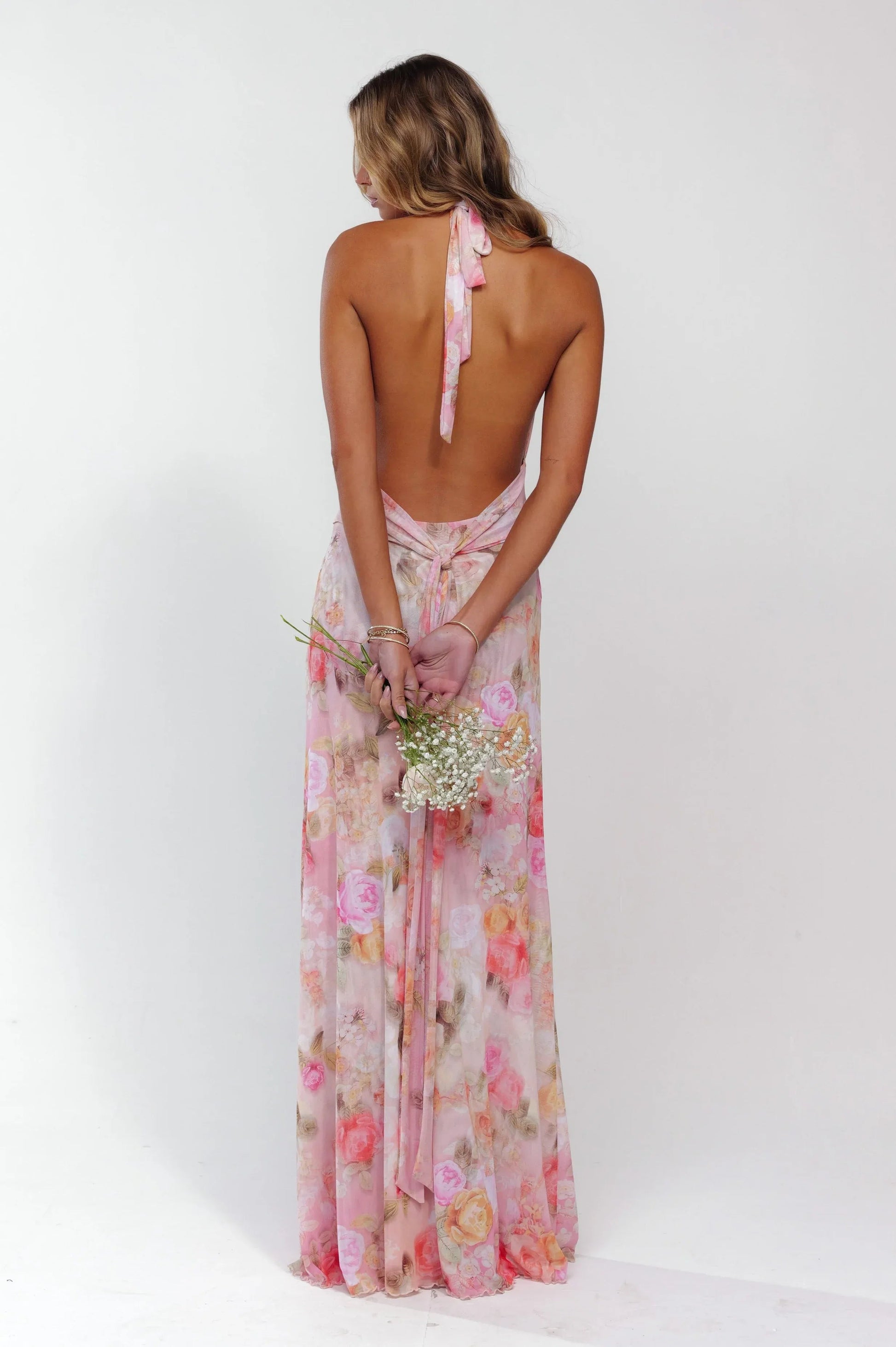 Back view of pink floral plunge maxi dress, showing open back and halter tie, holding flowers.