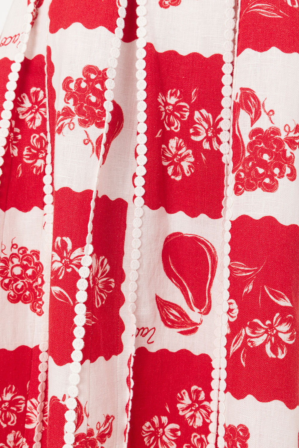 Close-up of the Savannah One-Shoulder Maxi Dress fabric, showing a vibrant red and white fruit and floral pattern with delicate white pom-pom trim detailing.