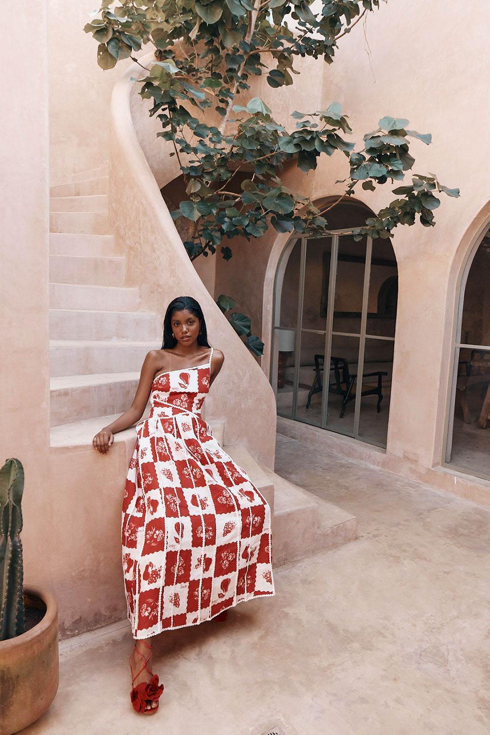 Savannah one-shoulder maxi dress in red and white checkered print, seated on steps, with designer shoes.