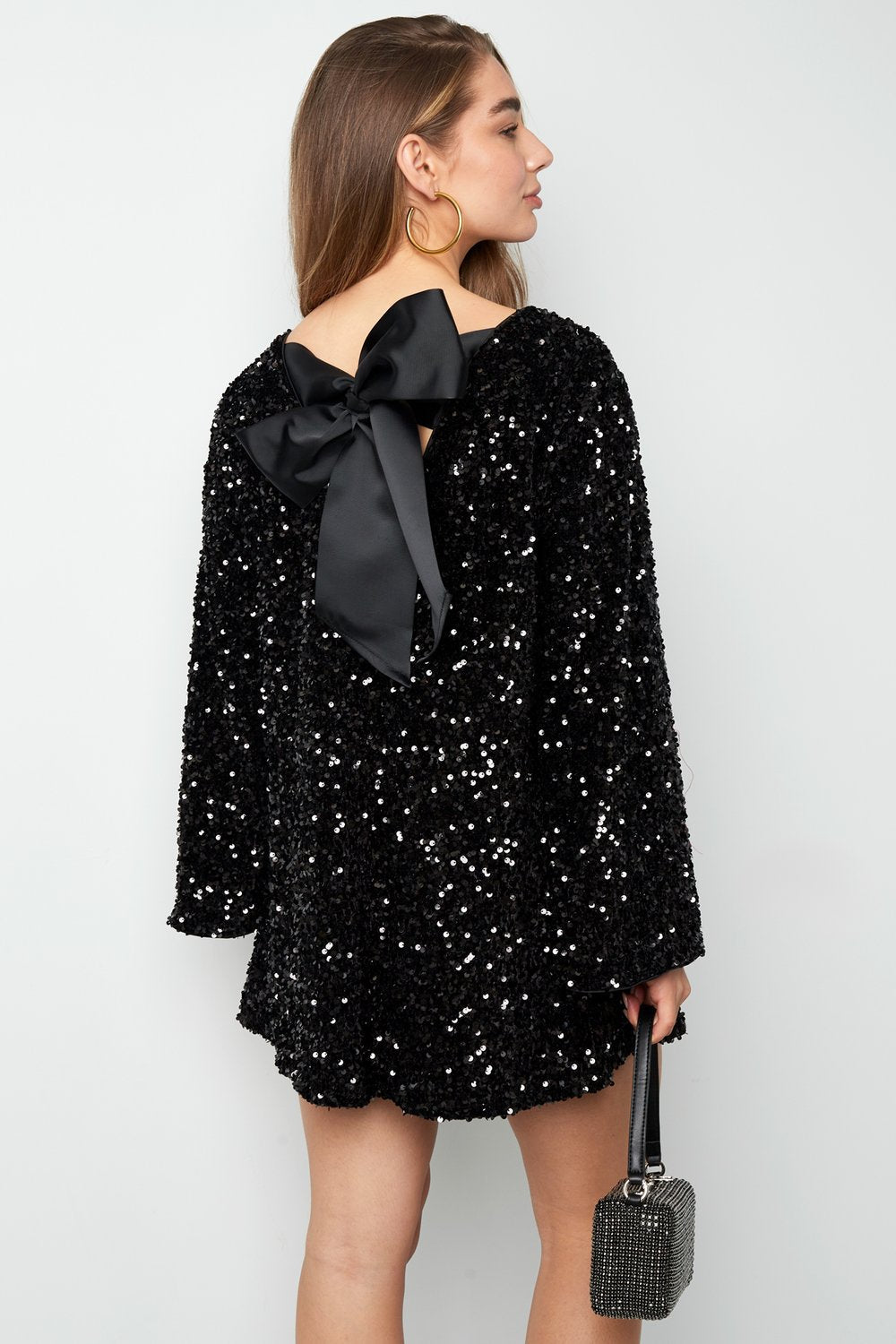 Back view of the Seraphina Bow Mini Dress in white, featuring a large black satin bow detail at the open back, long sleeves, and textured sequin fabric.