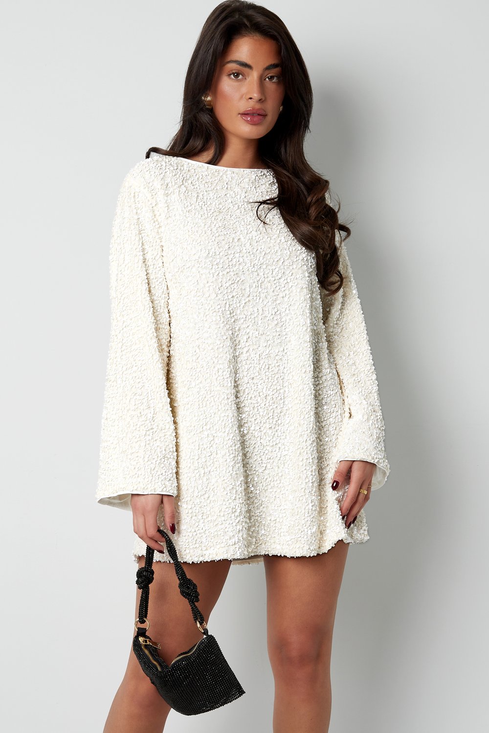 Front view of the white Seraphina Bow Mini Dress, showing the boat neck, long bell sleeves, and textured sequin fabric, paired with a small black beaded handbag.