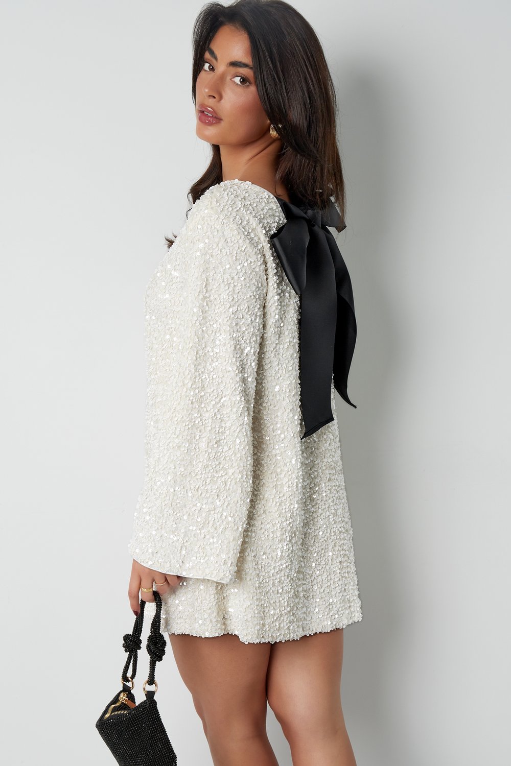 Side profile of the white Seraphina Bow Mini Dress, highlighting the black satin bow at the back, loose fit, and long sleeves, with a small black handbag.