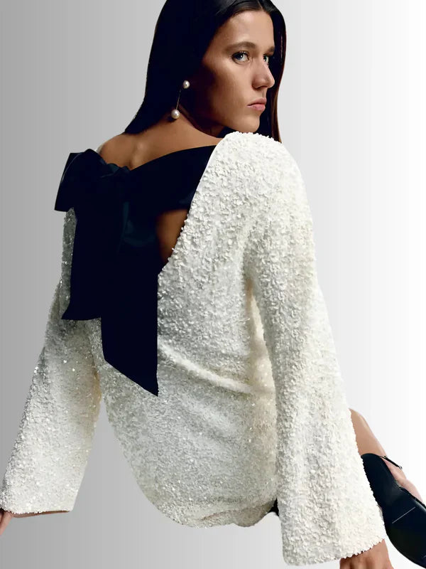 Dynamic back view of the white Seraphina Bow Mini Dress, emphasizing the deep open back and oversized black satin bow, with long textured sequin sleeves.