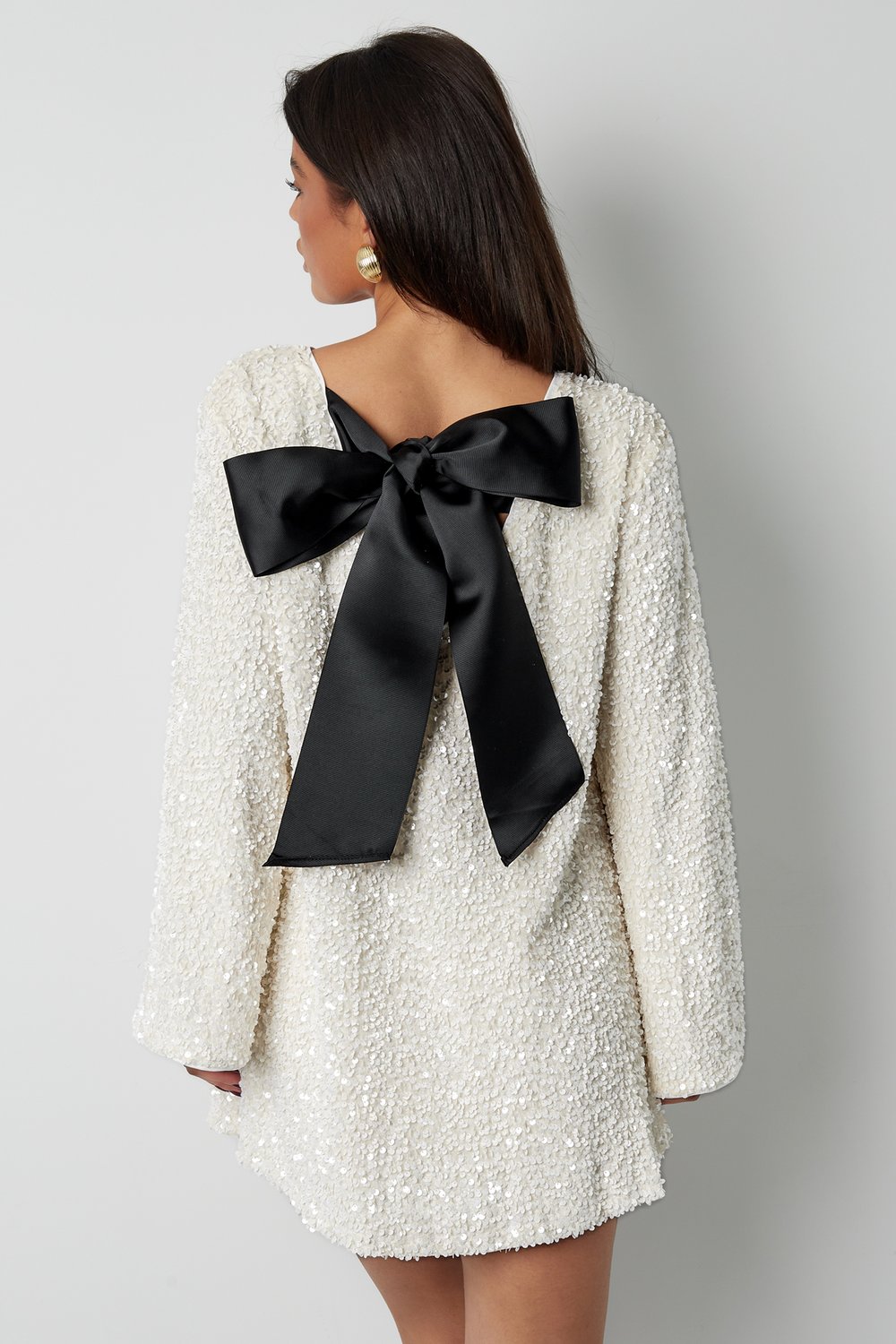 Back view of the Seraphina Bow Mini Dress in white, featuring a large black satin bow detail at the open back, long sleeves, and textured sequin fabric.