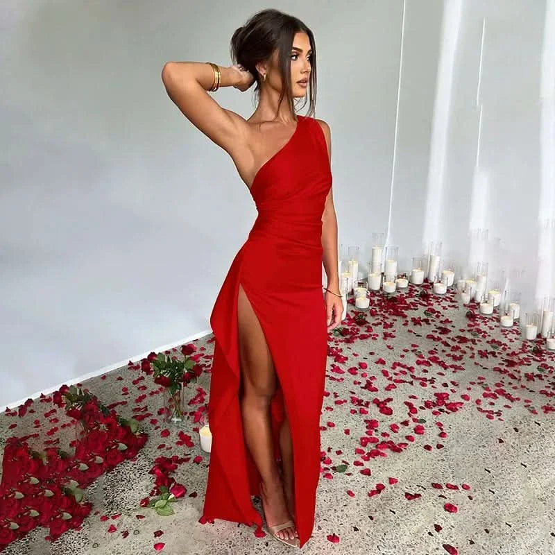 Side profile of a woman wearing a red one-shoulder Sienna Draped Maxi Dress with a high slit, surrounded by rose petals and candles.