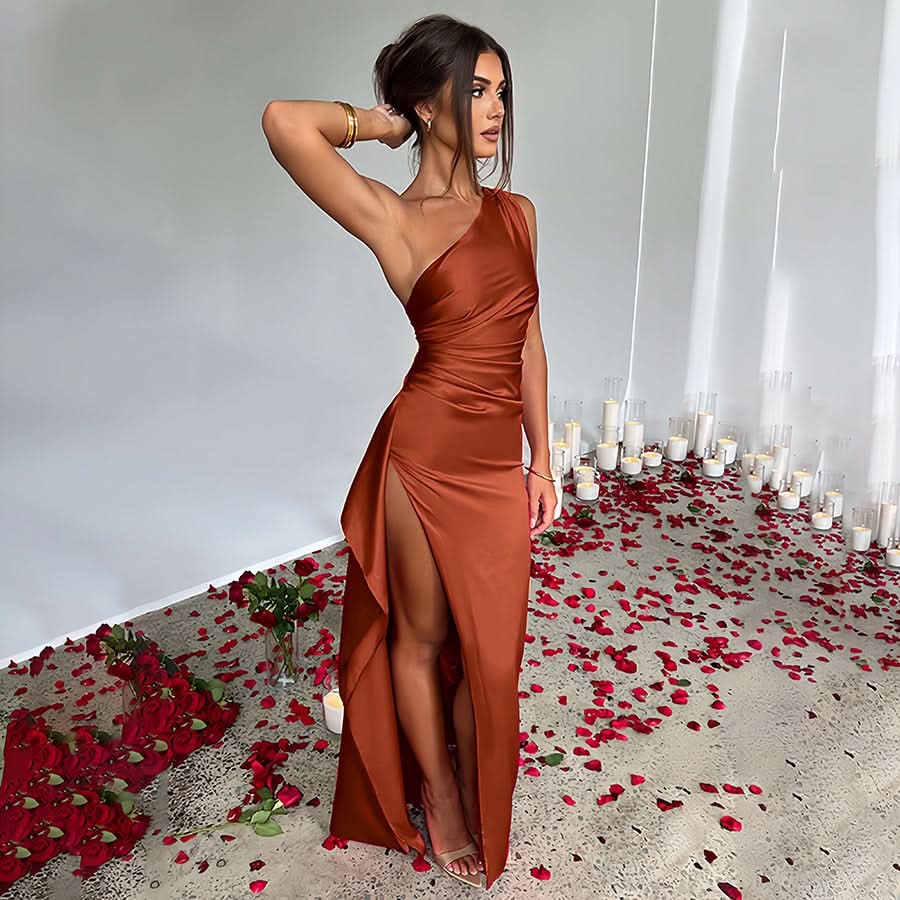Side profile of a woman in a rust-colored one-shoulder Sienna Draped Maxi Dress with a high slit, on a floor with rose petals.