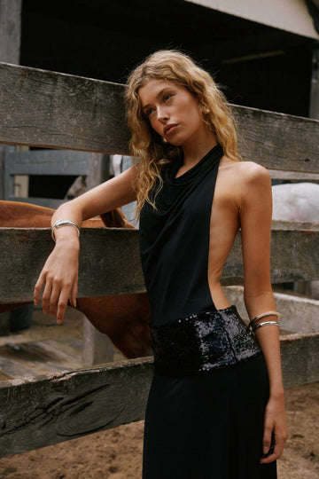 Black halter neck maxi dress with cowl neckline and sequin waist detail, leaning on a wooden fence.