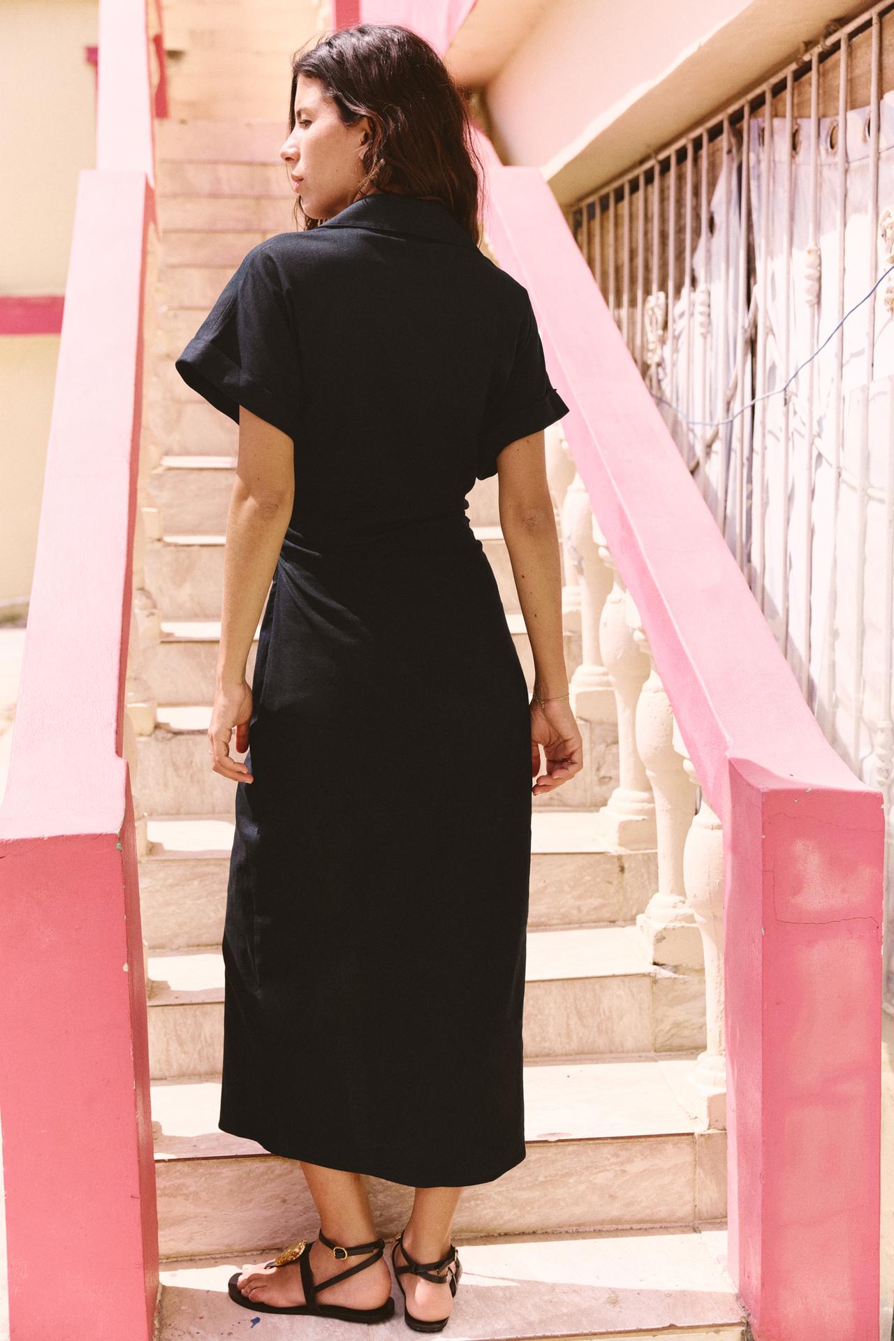 Back view of a woman wearing the black Sofia Button Midi Dress, walking up pink and white stairs, showcasing the full length of the dress and sandals.