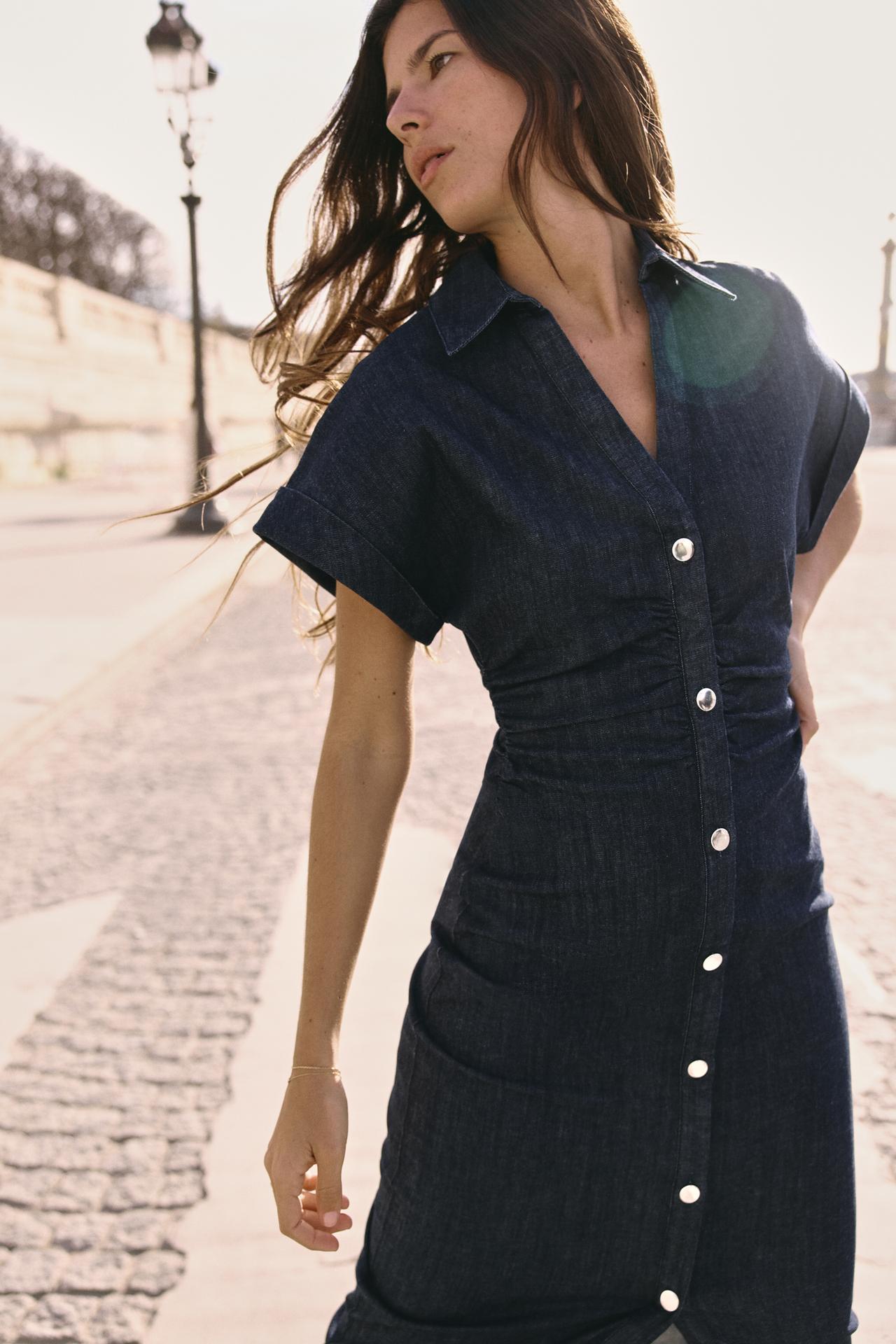 Side profile of the dark wash denim button-front midi dress, featuring rolled short sleeves and a cinched waist, with hair blowing.