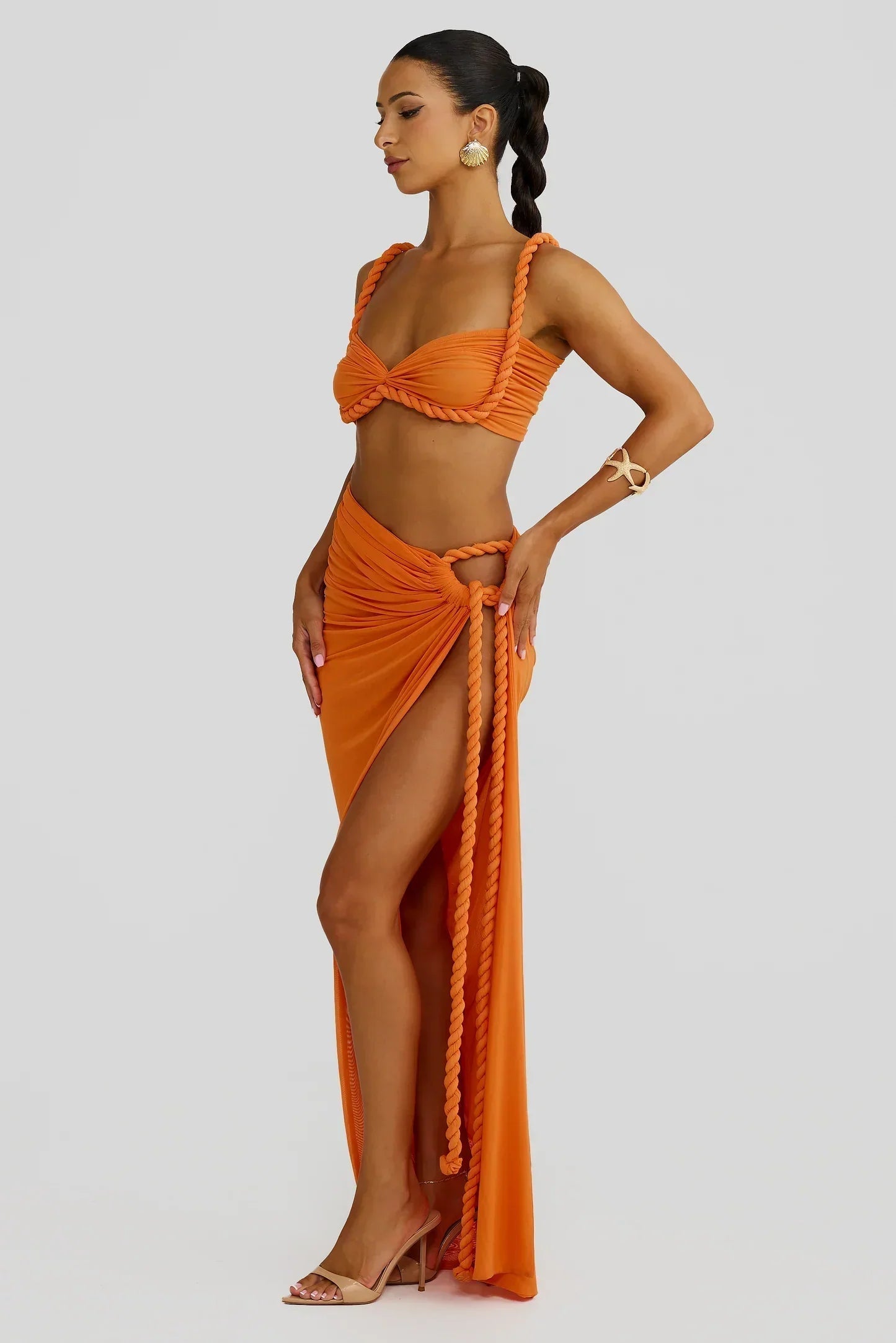 Side view of a woman in an orange braided maxi set, showcasing the bandeau top and the long, flowing skirt with a high slit and rope accents.