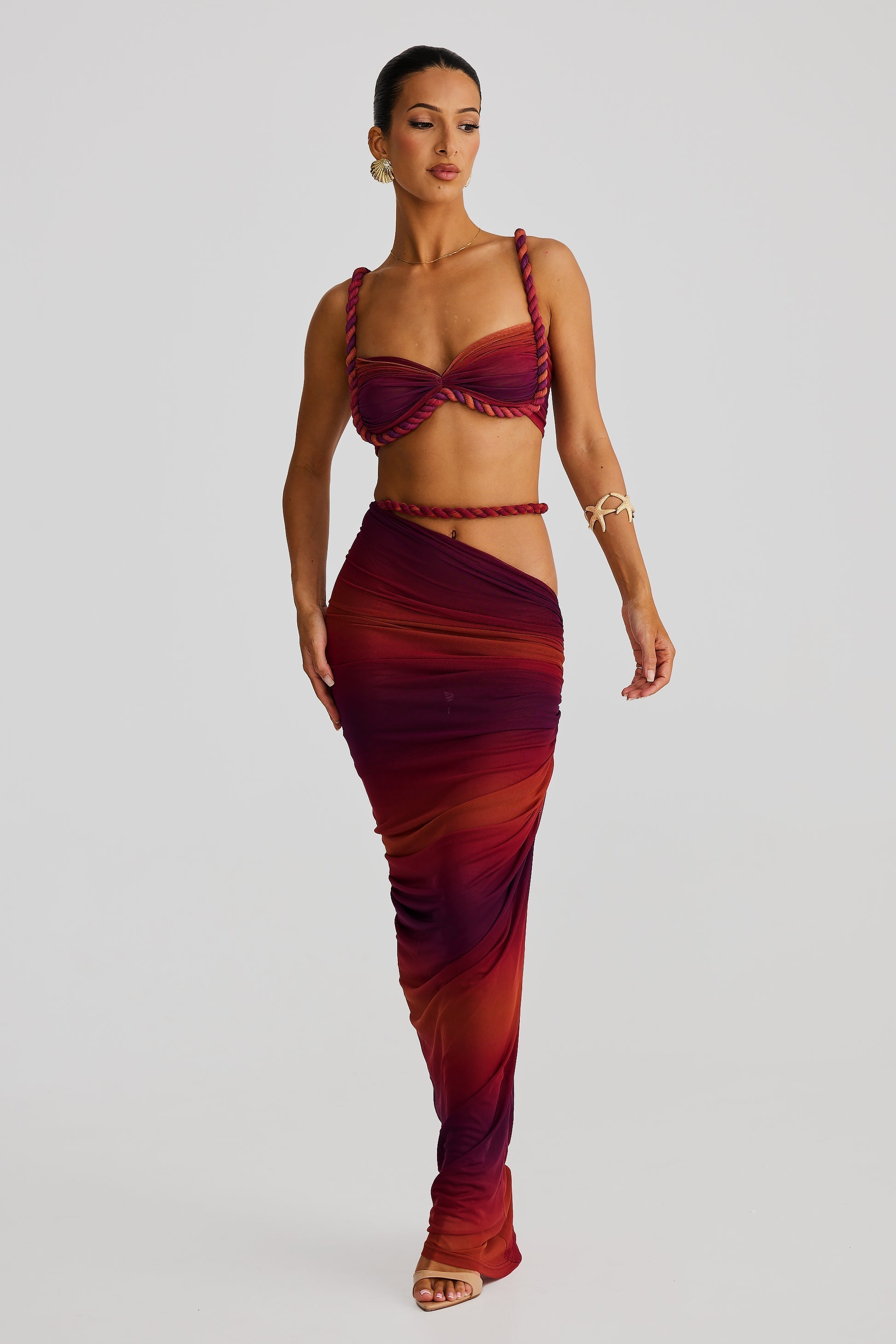 Full-length view of a woman in a vibrant ombre red and orange braided maxi dress set, with a bandeau top and a flowing draped skirt.
