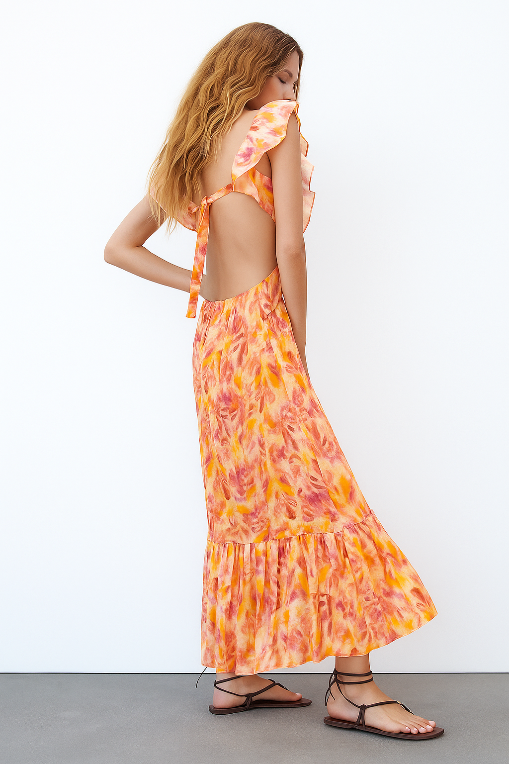 Side profile of a woman in an orange and pink floral maxi dress, showcasing the open back with tie detail and ruffled sleeves.