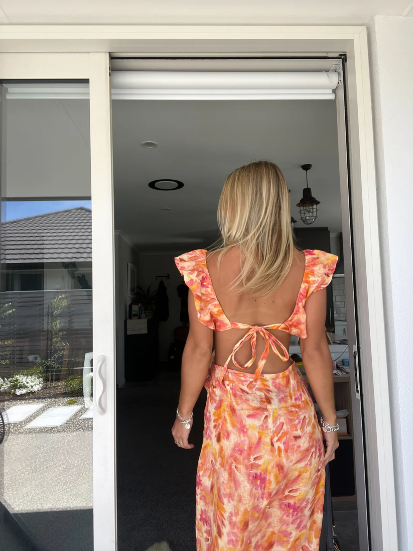 Back view of a woman walking away, showcasing the open back and tie detail of the orange and pink floral cutout maxi dress.