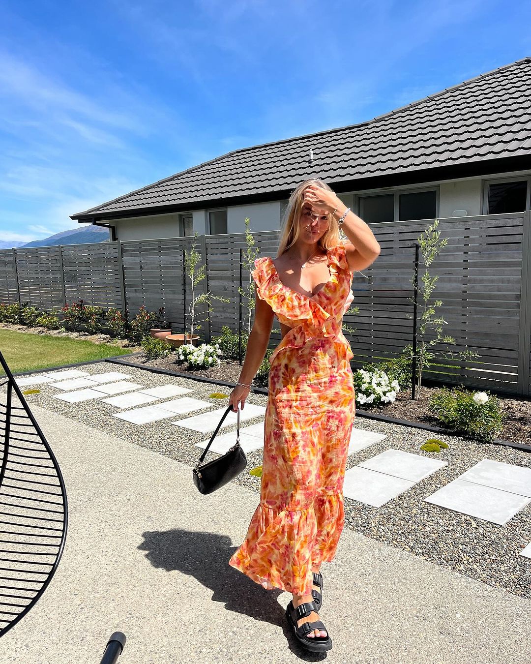 Full body shot of a woman in a Sunniva cutout maxi dress with a vibrant orange and pink floral print, holding a black handbag.